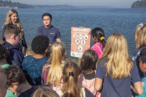 A group of school-aged children stand on the dock near the water.