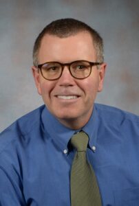 Man with glasses, short hair, blue shirt and grey tie