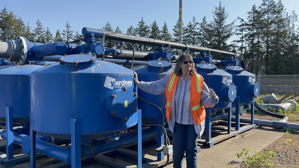 A woman with orange vest stands next to large blue tanks during a tour of the Port of Olympia stormwater treatment facility.